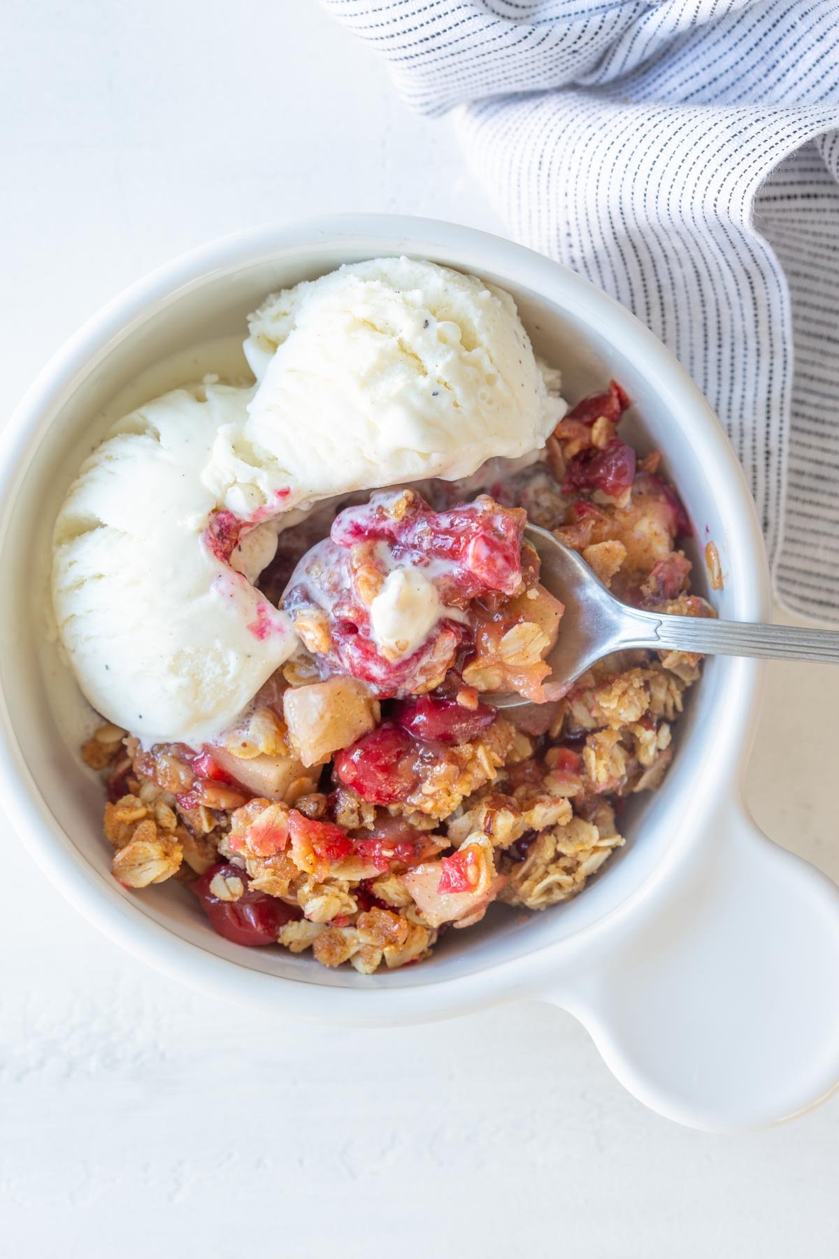 A spoon in a white bowl of cranberry apple crisp with vanilla ice cream.