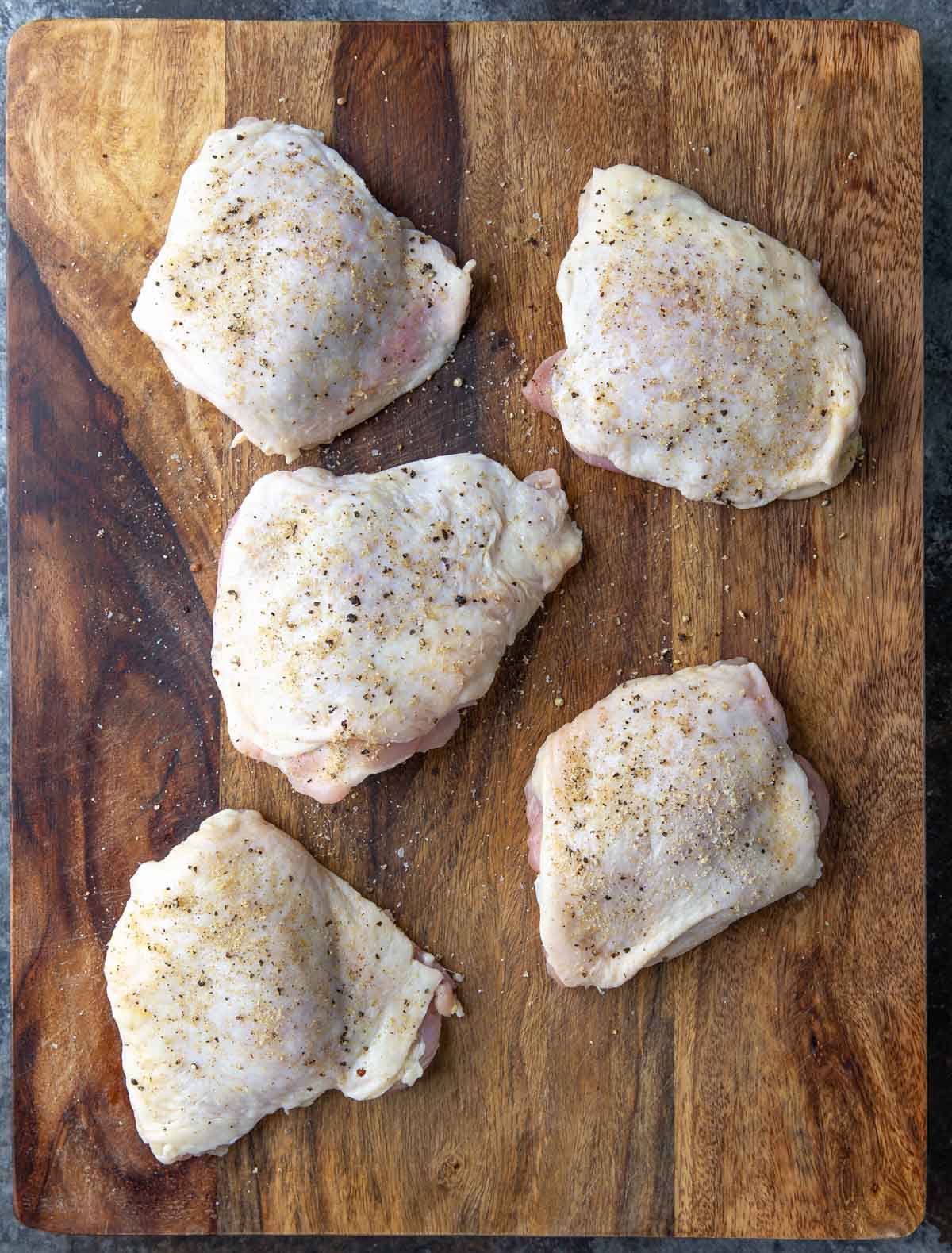 Overhead view of five raw chicken thighs that have been seasoned on a cutting board.