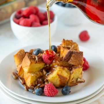 Syrup being poured over baked eggnog French toast with fruit in a white plate.