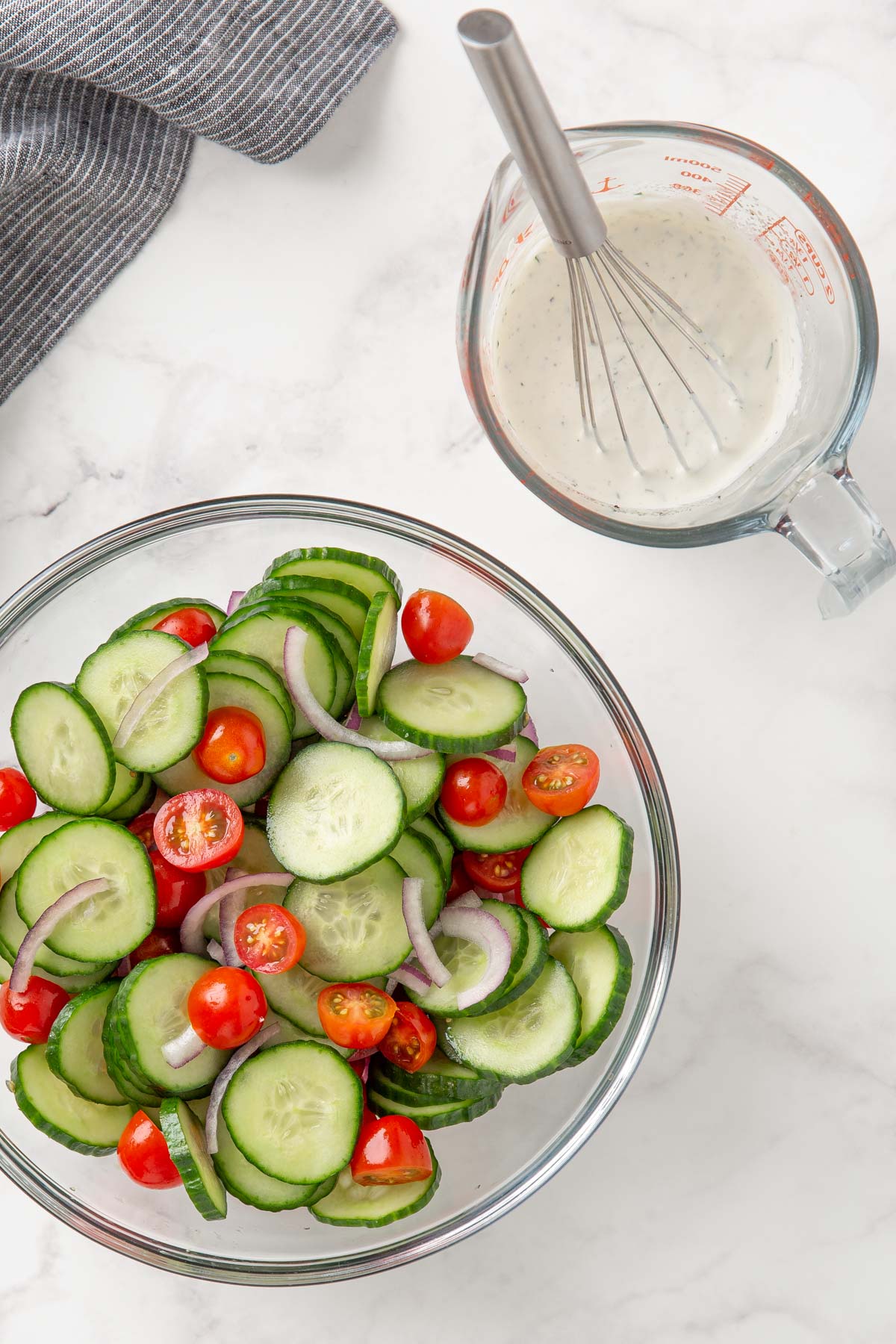 Cucumbers, tomato and onion in a bowl beside a measuring cup of creamy dressing.