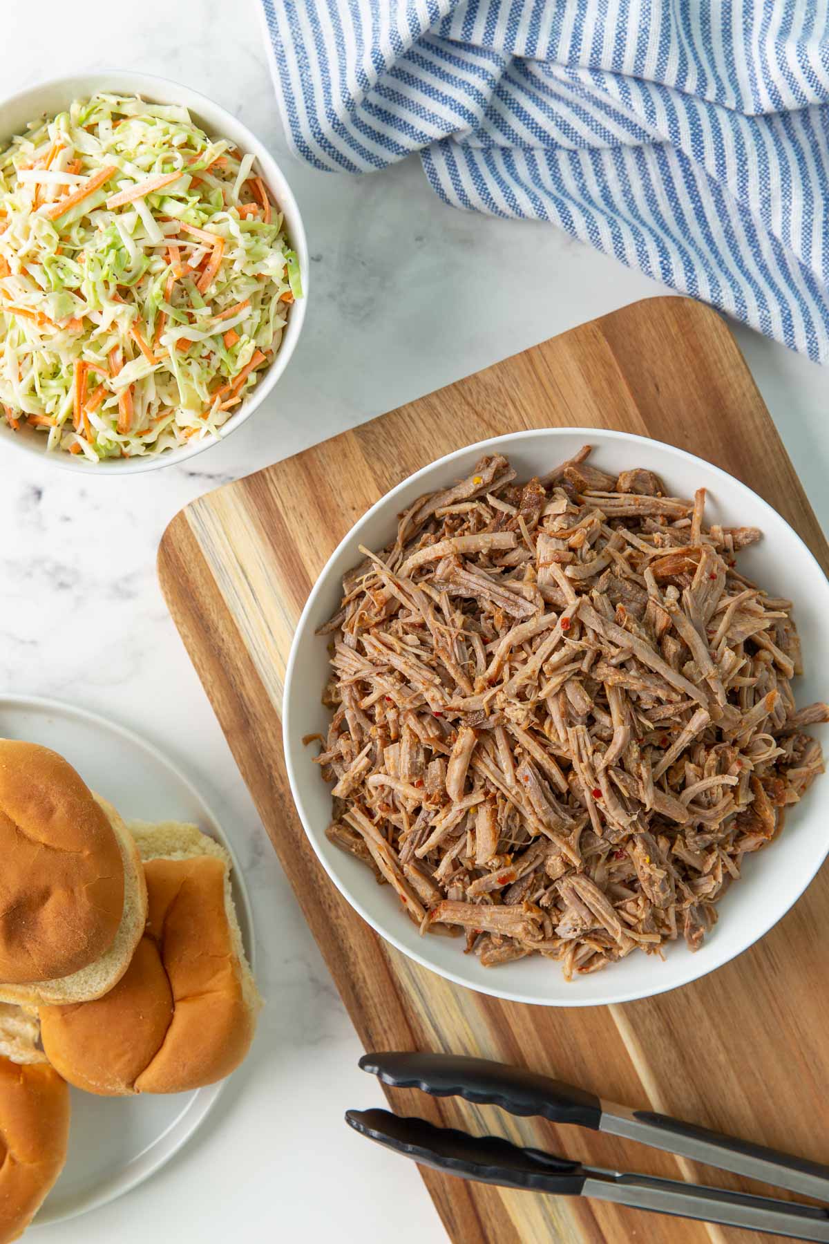 A bowl of pulled pork, a bowl of coleslaw and a plate with sandwich buns.