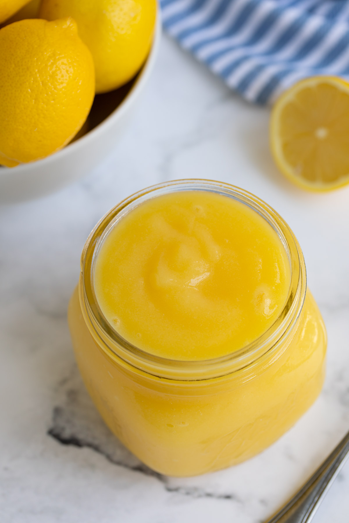 An open jar of microwave lemon curd on a white surface by a bowl of lemons.