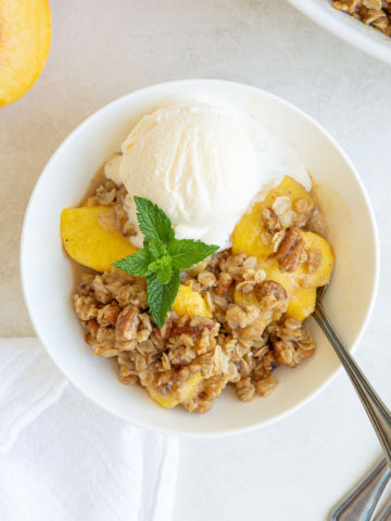 Peach crisp in a white bowl with a scoop of ice cream and a mint sprig.
