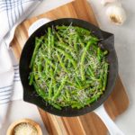 Garlic parmesan green beans in a skillet on a cutting board by a kitchen towel.