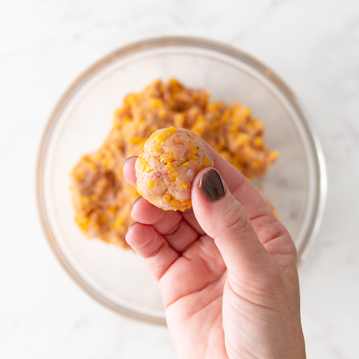 A hand holding a formed sausage cheese ball over a bowl of mixture.