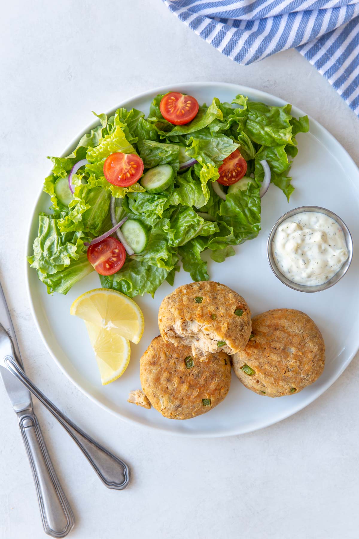 Three salmon patties on a white plate with a salad, lemon wedges and tartar sauce.