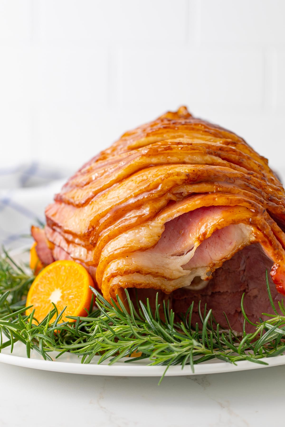 Front view of a glazed spiral ham on a white platter with rosemary and an orange.