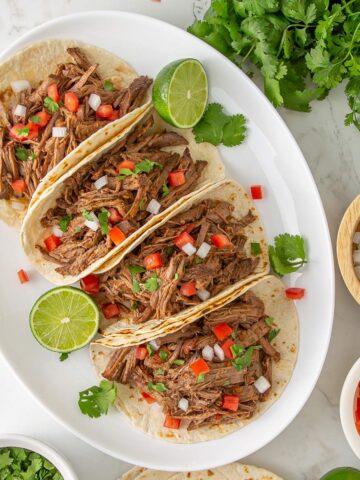 Four slow cooker Mexican shredded beef soft tacos on a white platter.