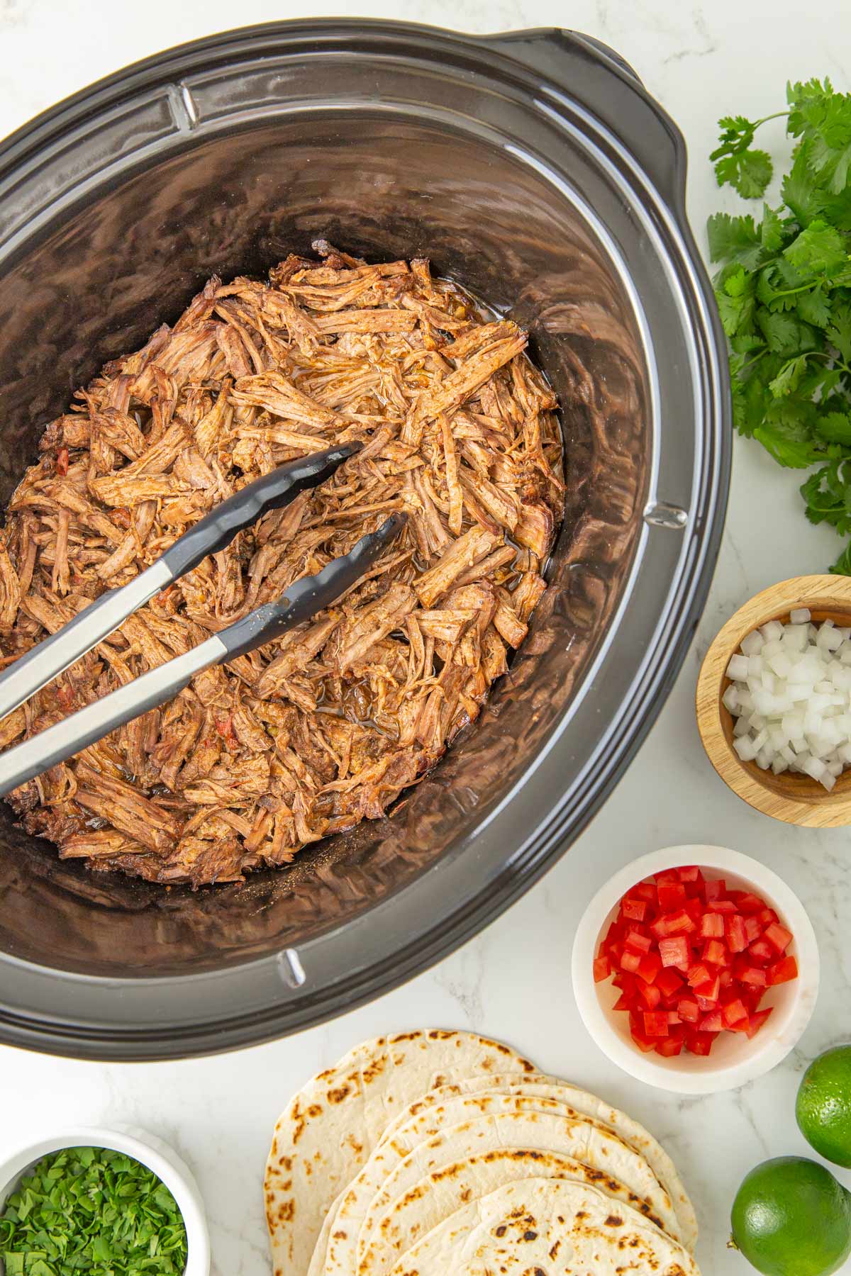 Mexican shredded beef in a slow cooker with a set of tongs beside bowls of taco toppings.