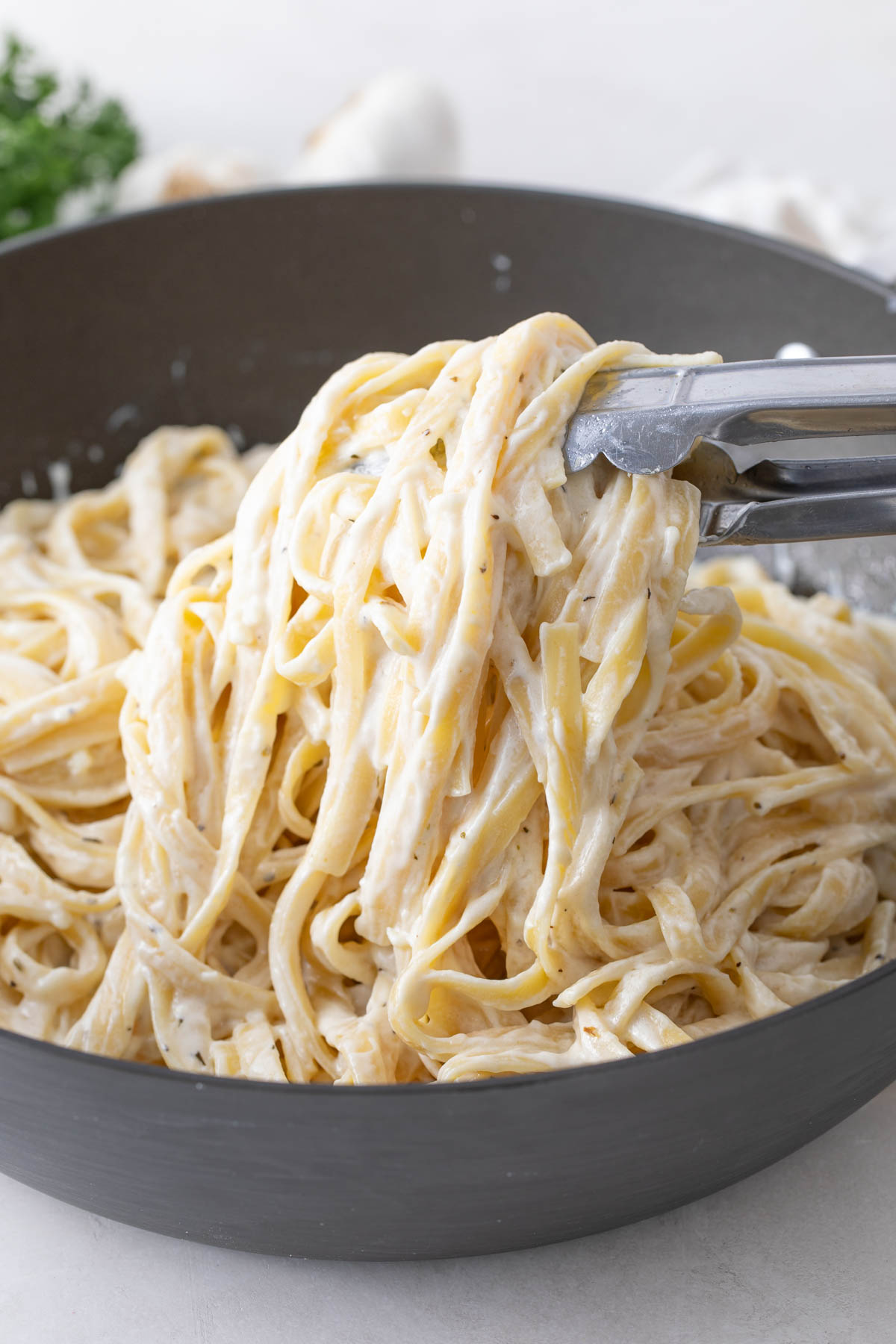 Front view of a pair of tongs holding fettucine with Alfredo sauce in a pan.