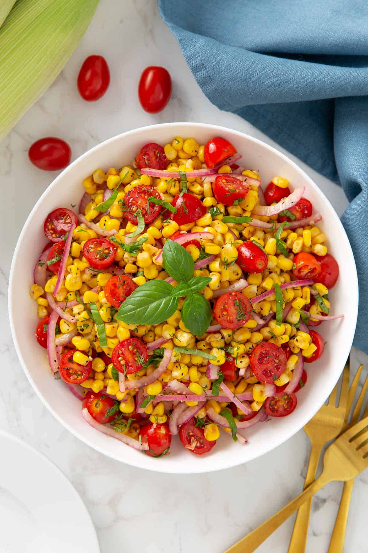 Overhead view of corn and tomato salad in a white bowl beside a blue napkin.