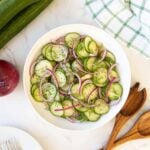 Cucumber onion salad in a white bowl beside wooden salad servers.