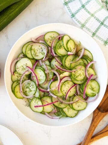 Cucumber onion salad in a white bowl beside wooden salad servers.