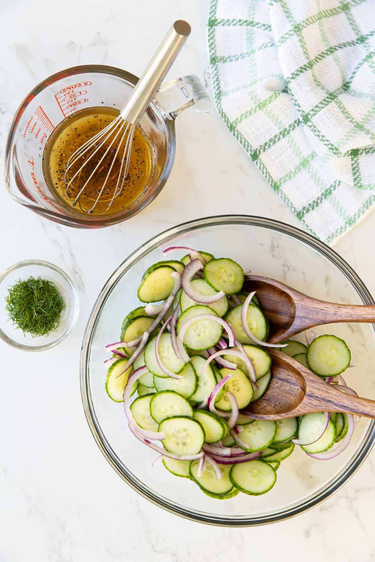 A bowl of cucumbers and onions beside a small bowl of dill and a measuring cup of vinegar mixture.