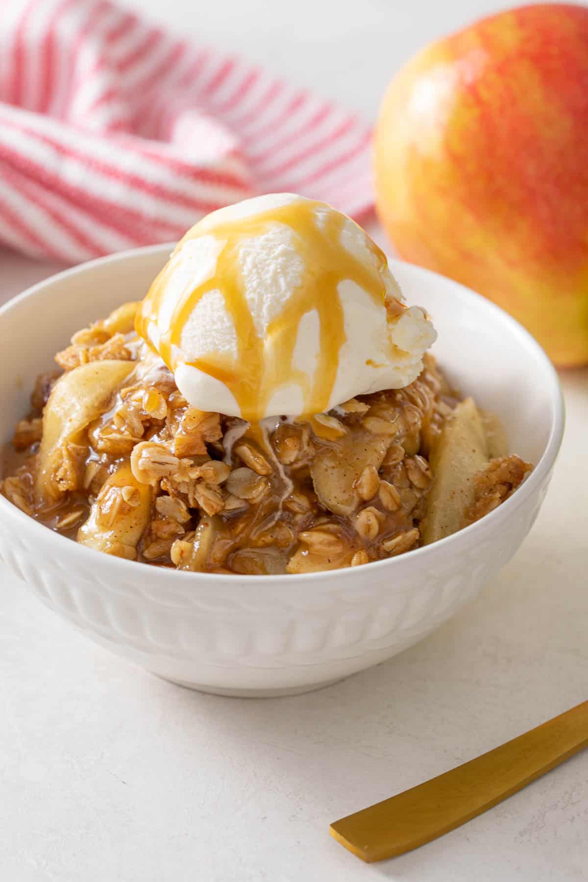 Apple crisp topped with a scoop of ice cream and drizzled with caramel sauce in a white bowl.