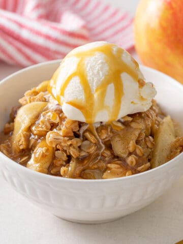 A white bowl of apple crisp topped with a scoop of vanilla ice cream and caramel sauce.