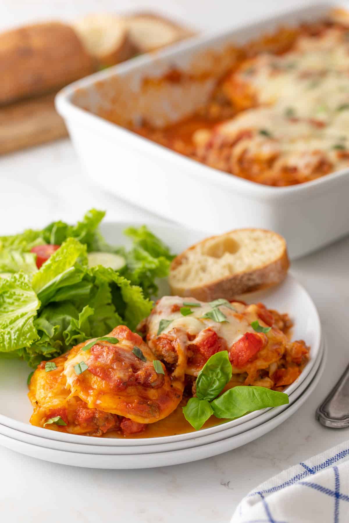 A serving of baked ravioli on a white plate with salad and a slice of French bread.
