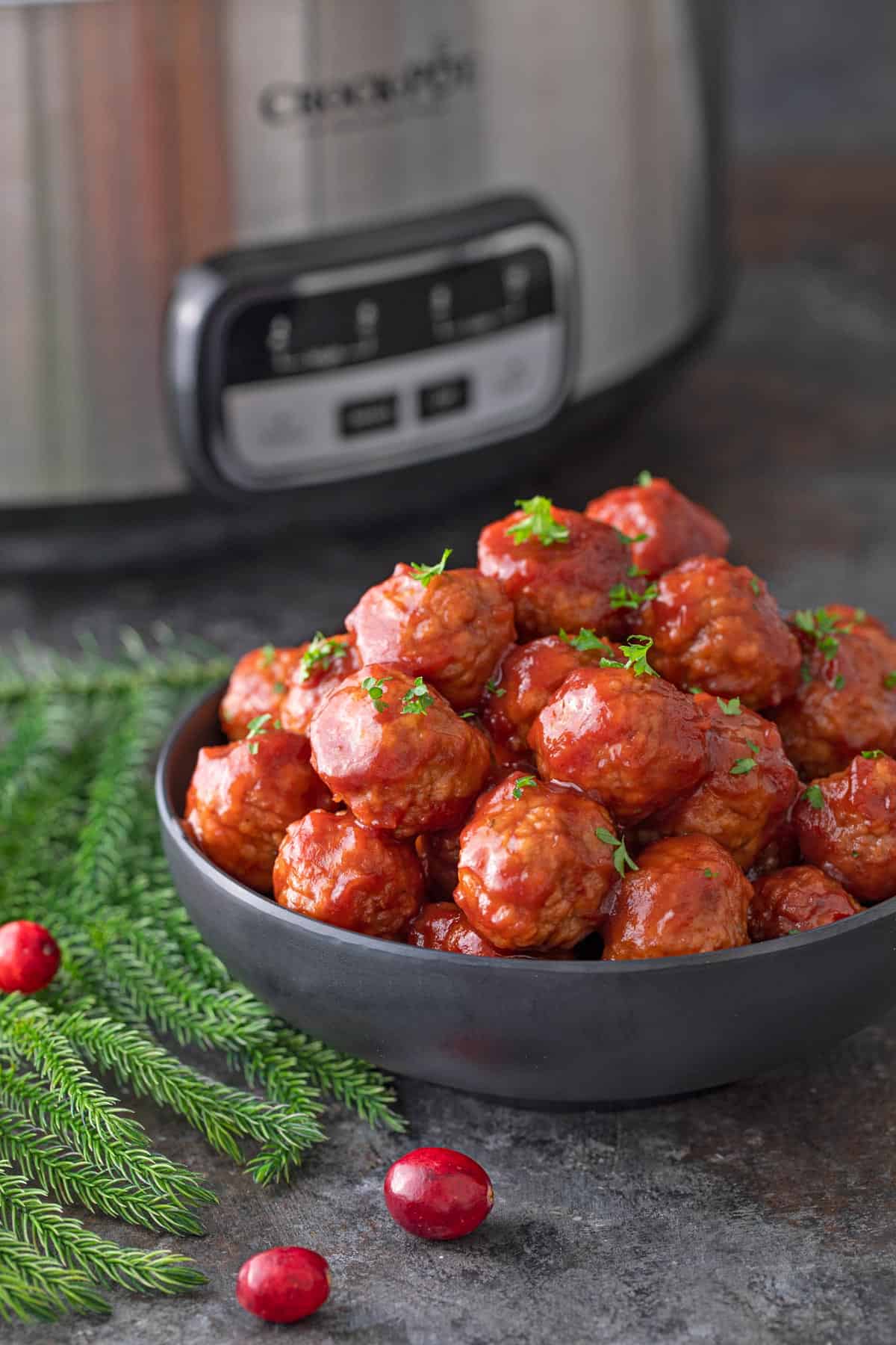 A black bowl of cranberry meatballs with a crock pot in the background.