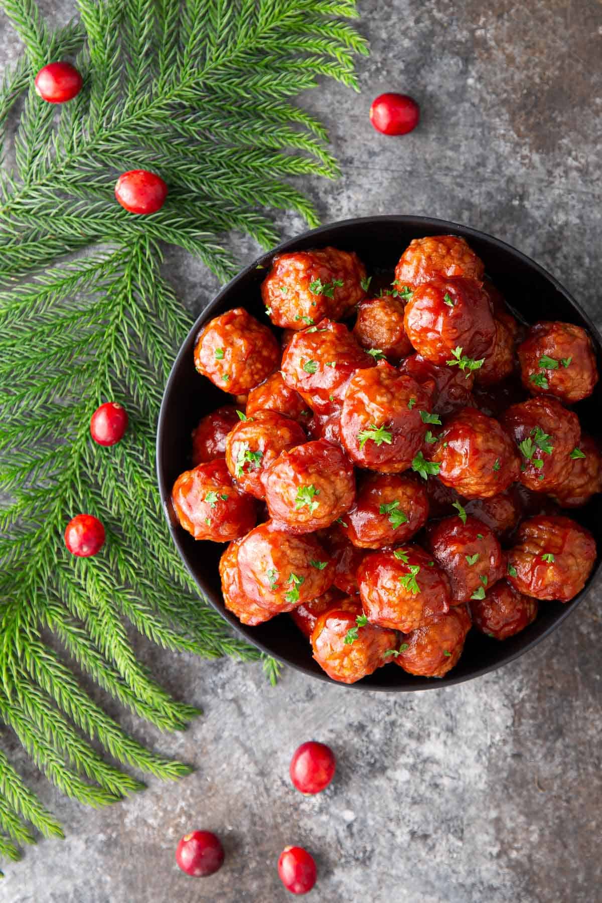 Overhead view of cranberry meatballs in a black bowl beside festive greenery and fresh cranberries.