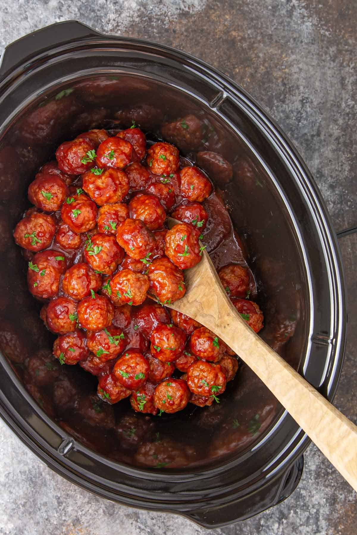 Cranberry meatballs in an oval crockpot with a wooden spoon.