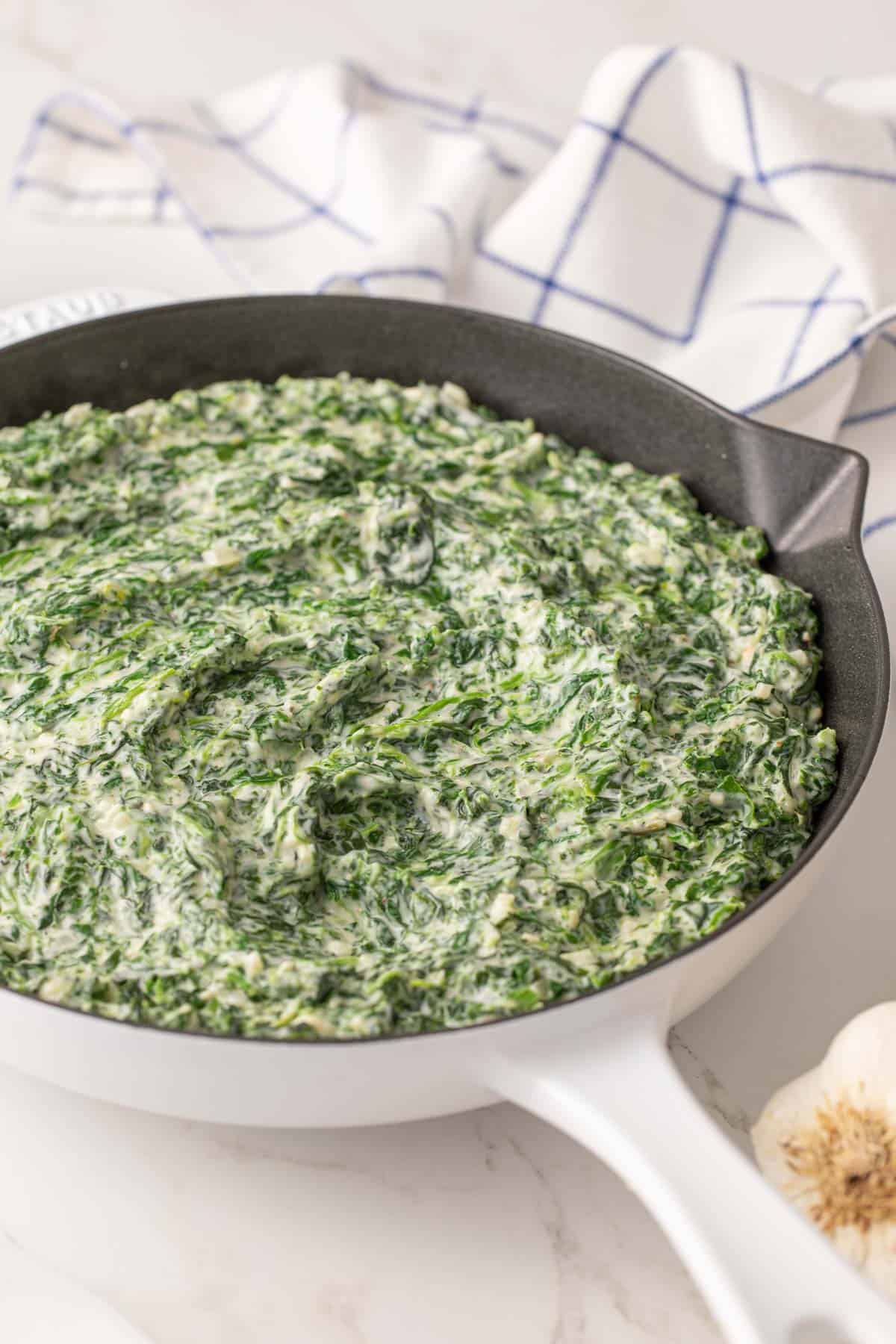 Closeup view of easy creamed spinach in a white skillet by a white and blue dish towel.