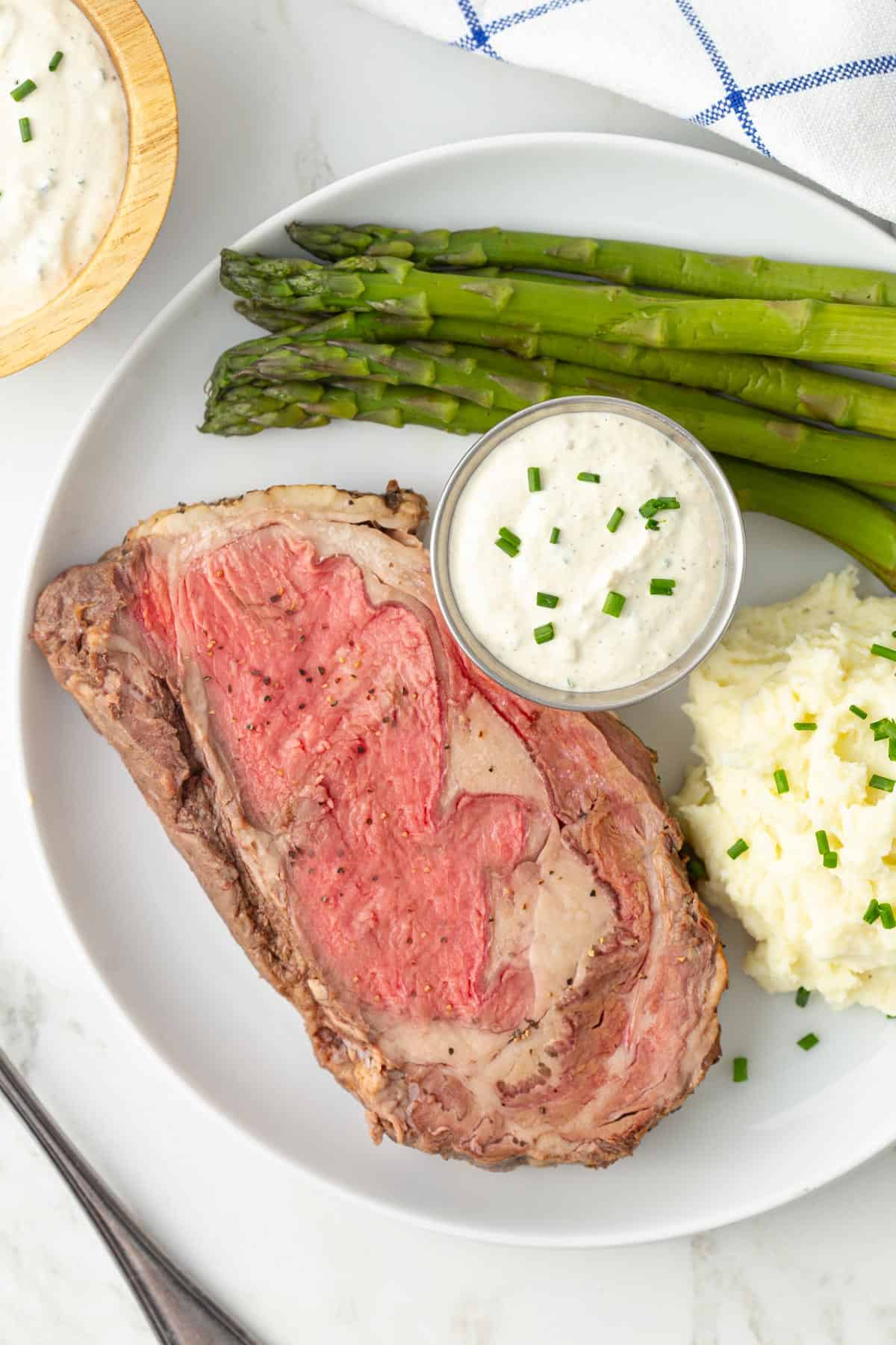 A slice of prime rib, asparagus, mashed potatoes and a condiment cup of horseradish sauce on a plate.