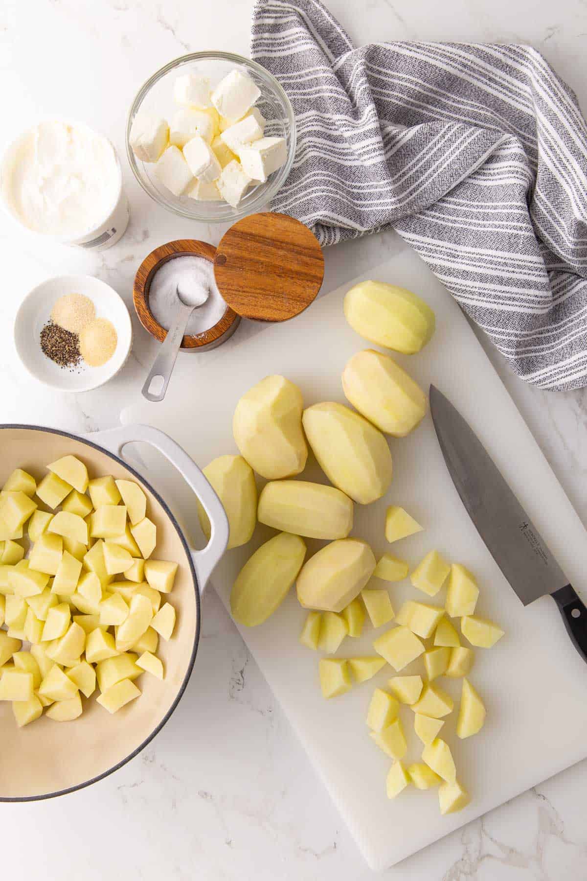 Overhead view of peeled potatoes on a cutting board, seasonings, cubed cream cheese and sour cream.