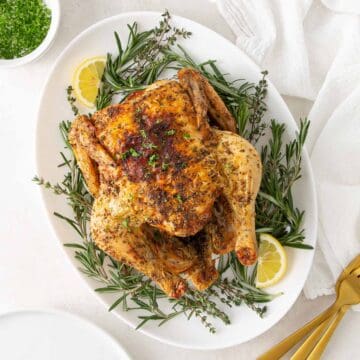 Air fryer whole chicken on a white platter surrounded by fresh rosemary and thyme.