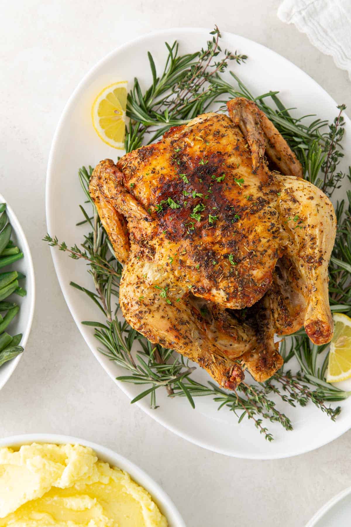 Air fryer whole chicken on a platter beside a bowl of green beans and a bowl of mashed potatoes.