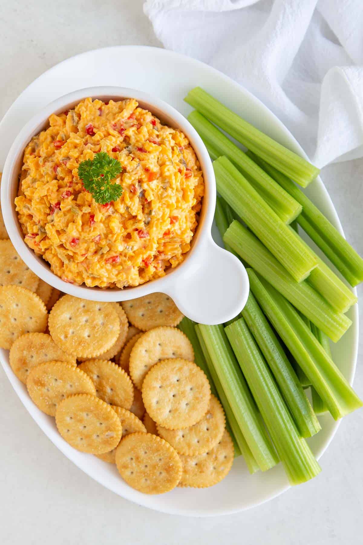 Overhead view of a bowl of pimento cheese spread on an oval white platter with crackers and celery sticks.