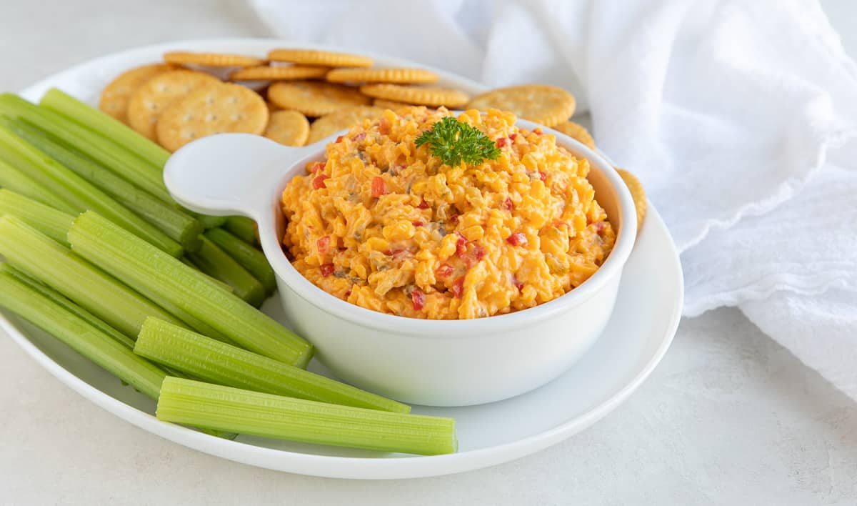 A bowl of pimento cheese spread on an oval platter with crackers and celery sticks.