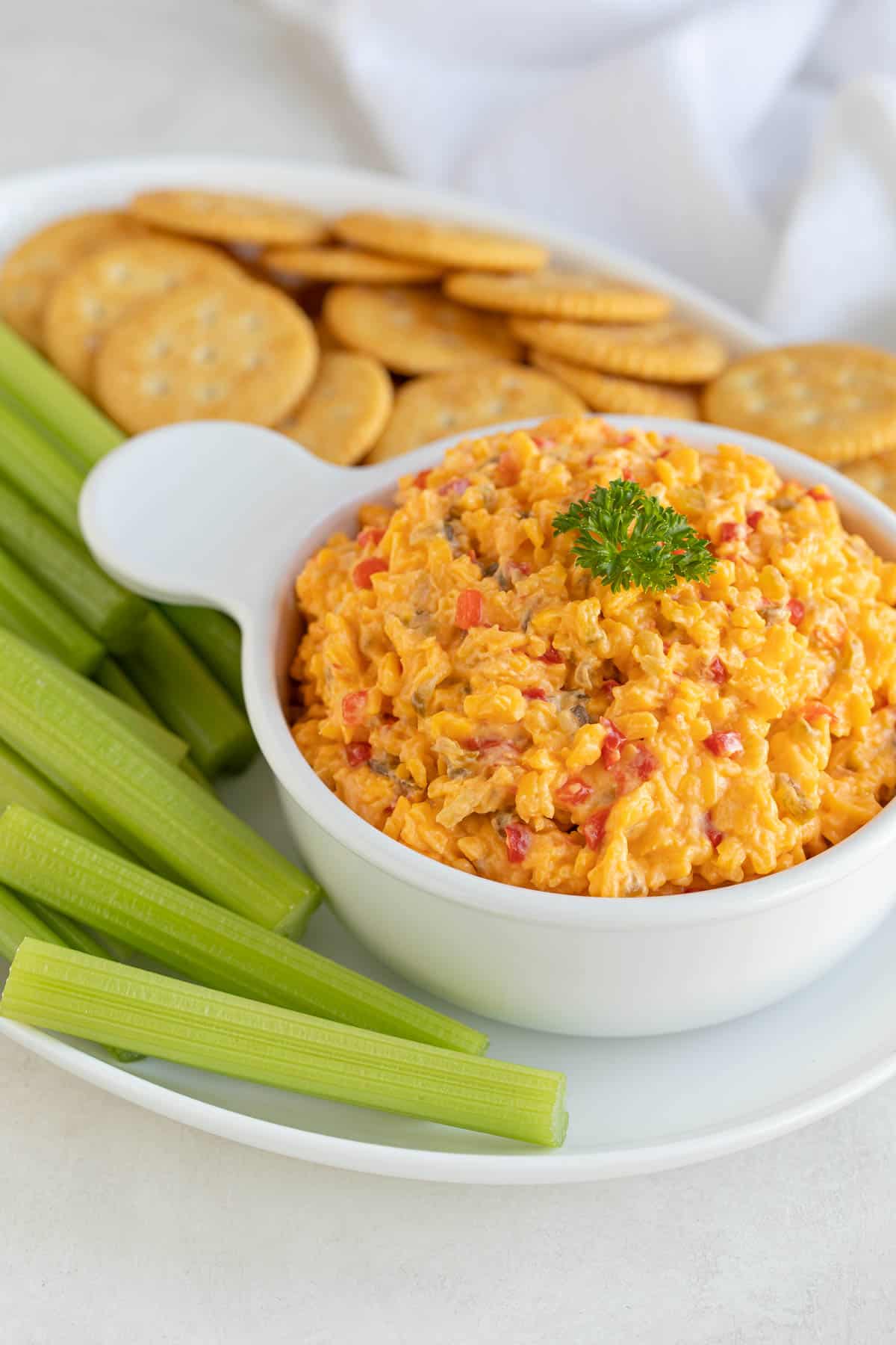 Front view of pimento cheese spread in a white bowl on a platter with crackers and celery sticks.