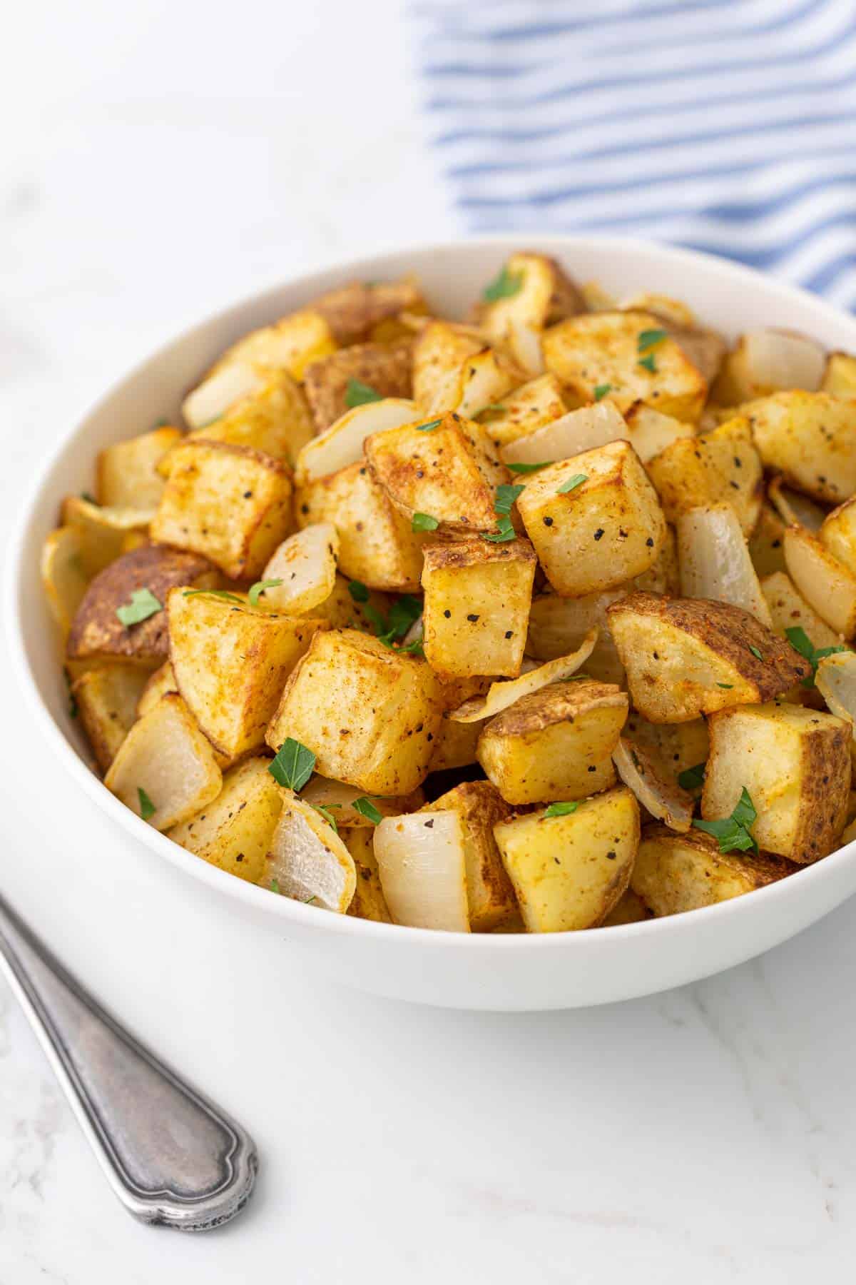 Air fried potatoes and onions in a white bowl by a serving spoon.