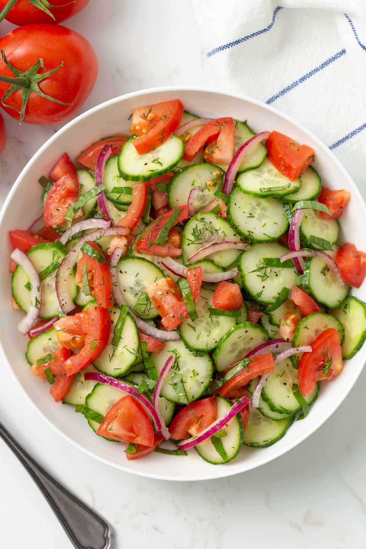 Overhead view of cucumber tomato salad in a white bowl by a blue striped kitchen towel.