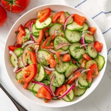 Overhead view of cucumber tomato salad in a white bowl by a serving spoon.