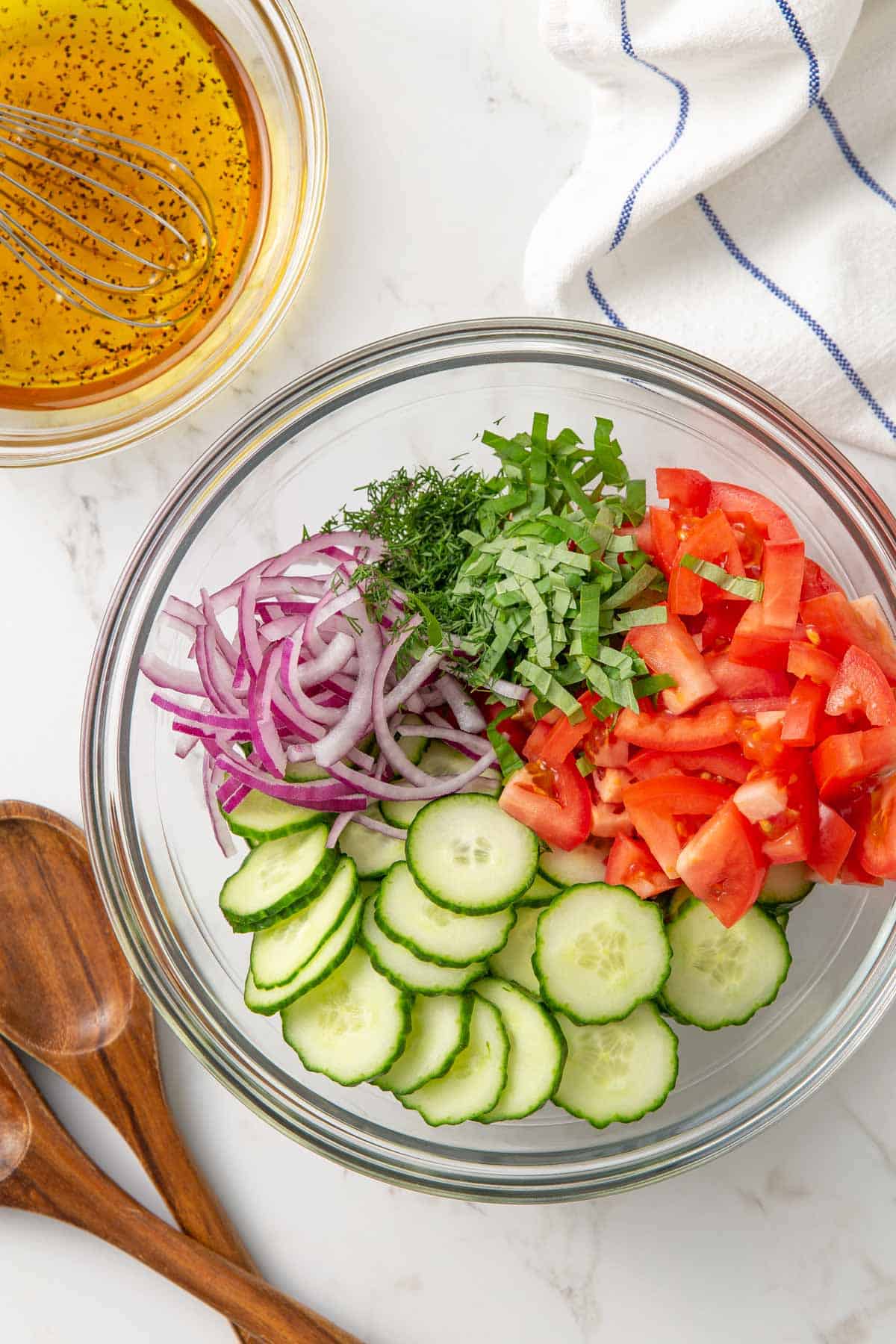 Cucumbers, tomatoes, red onion, dill and basil in a bowl by a bowl of vinaigrette dressing.