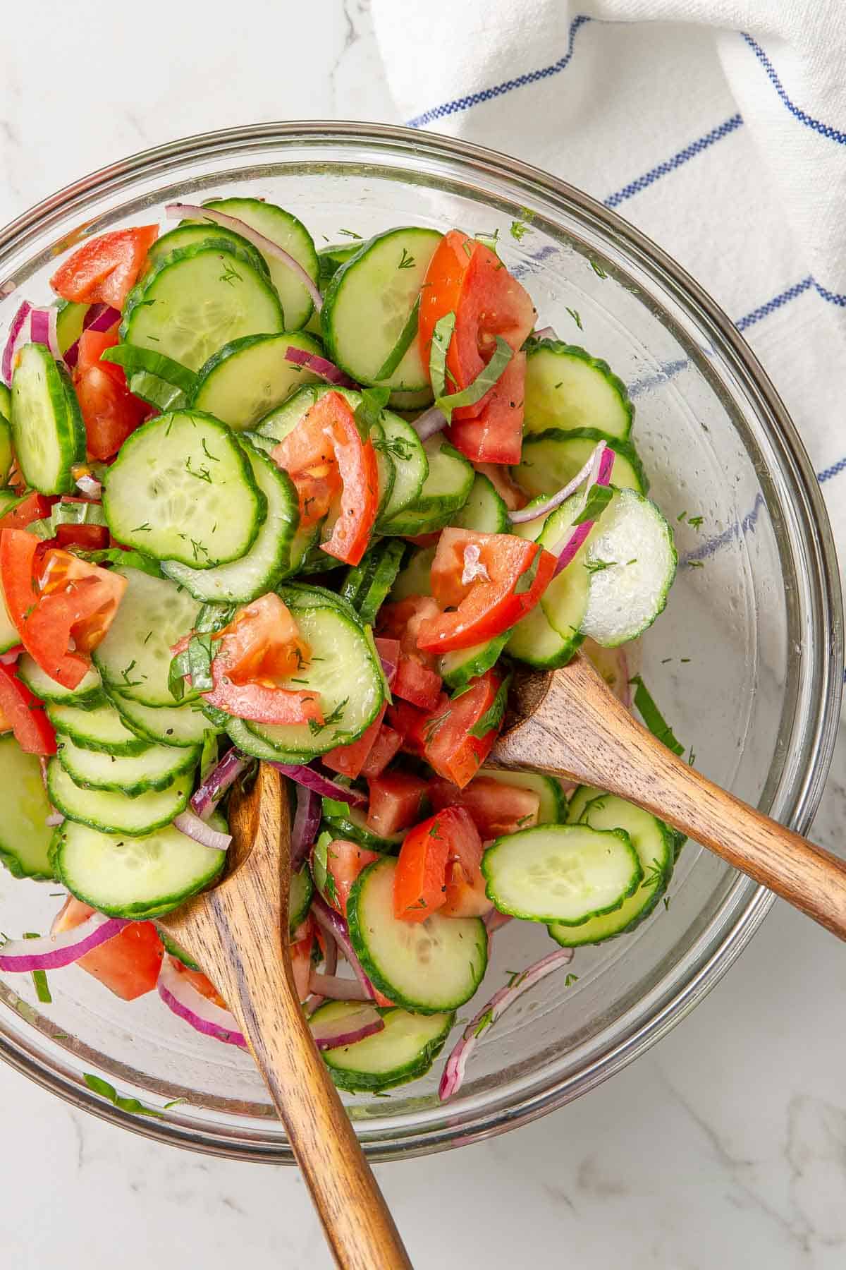 A bowl of cucumber tomato salad with wooden salad tongs.