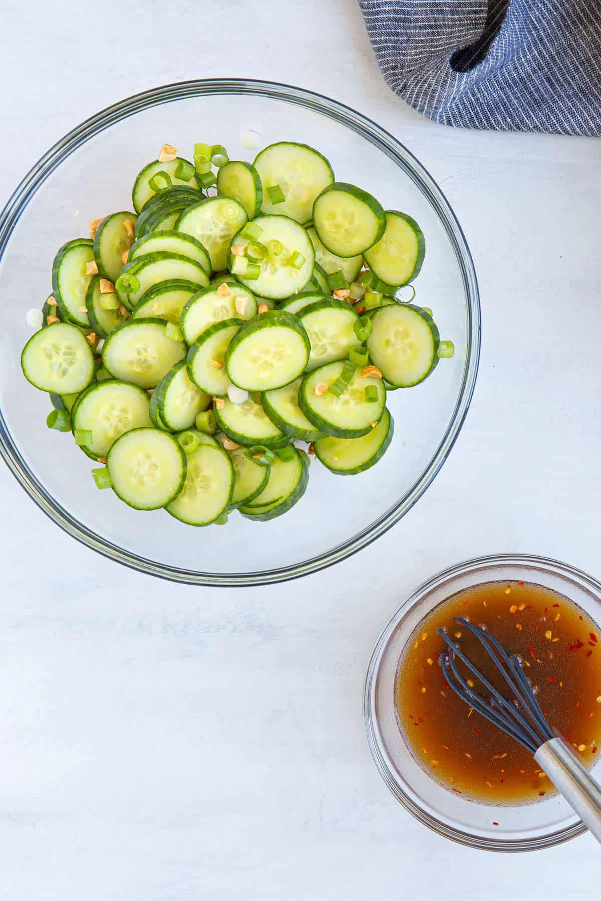 Overhead view of a bowl of sliced cucumbers, onions and peanuts beside a bowl of Thai dressing.