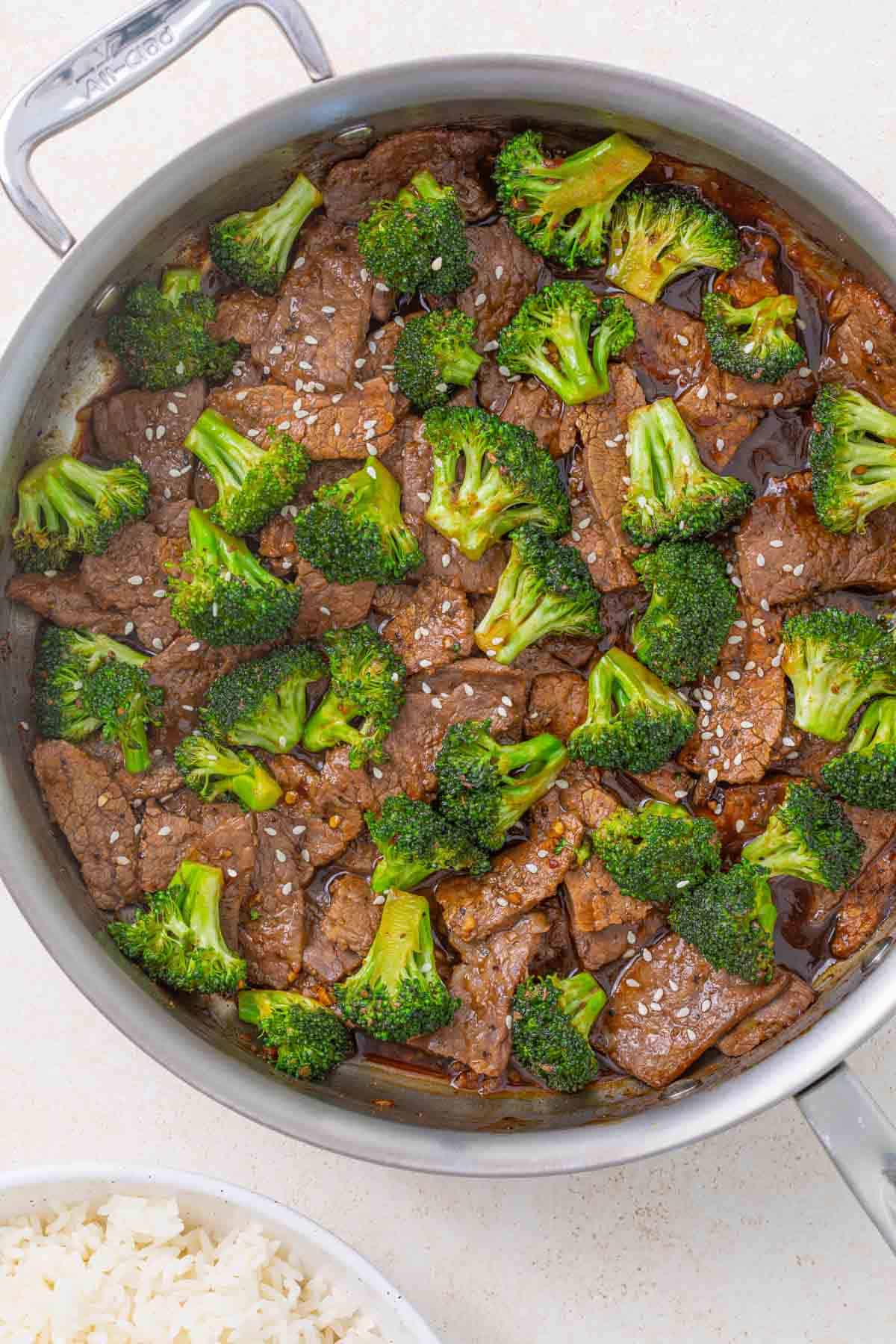 Beef and broccoli stir-fry in a sauté pan beside a bowl of white rice.
