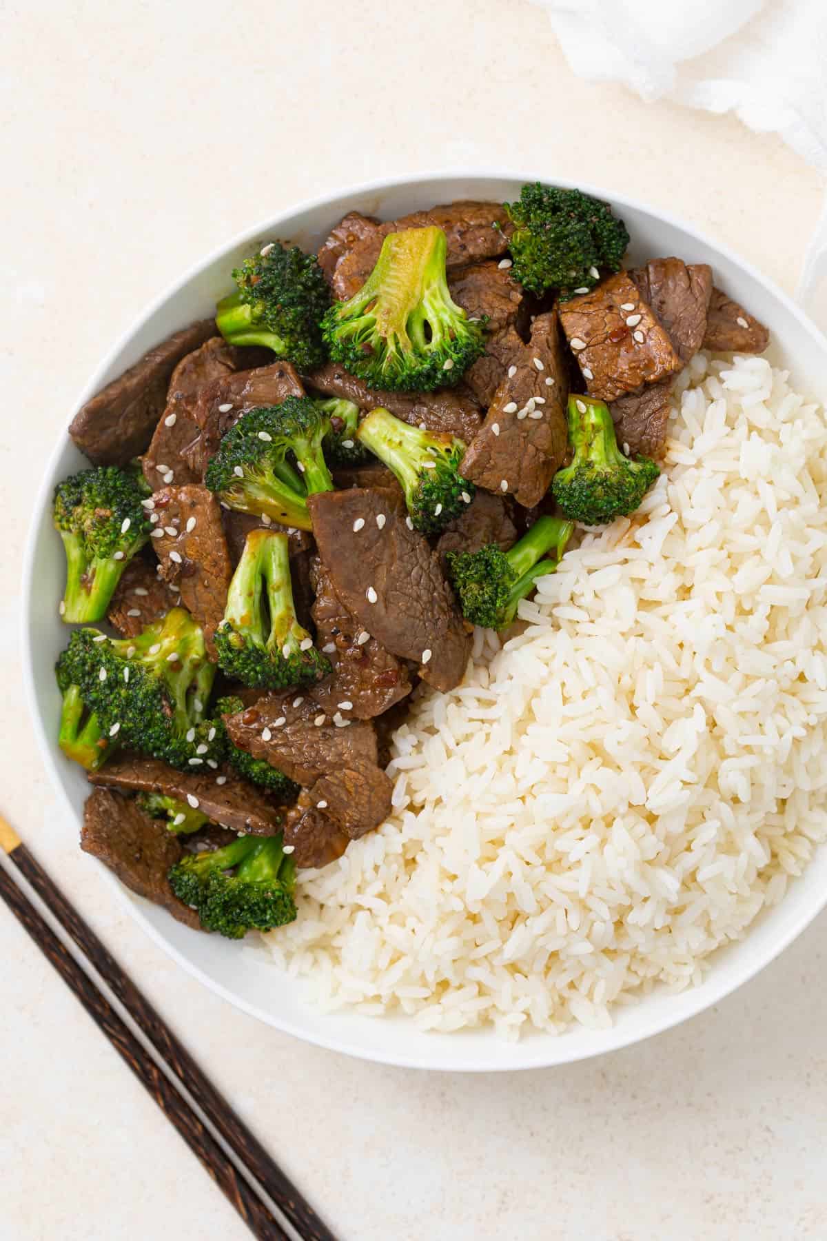 Beef and broccoli in a bowl with white rice beside a set of chopsticks.