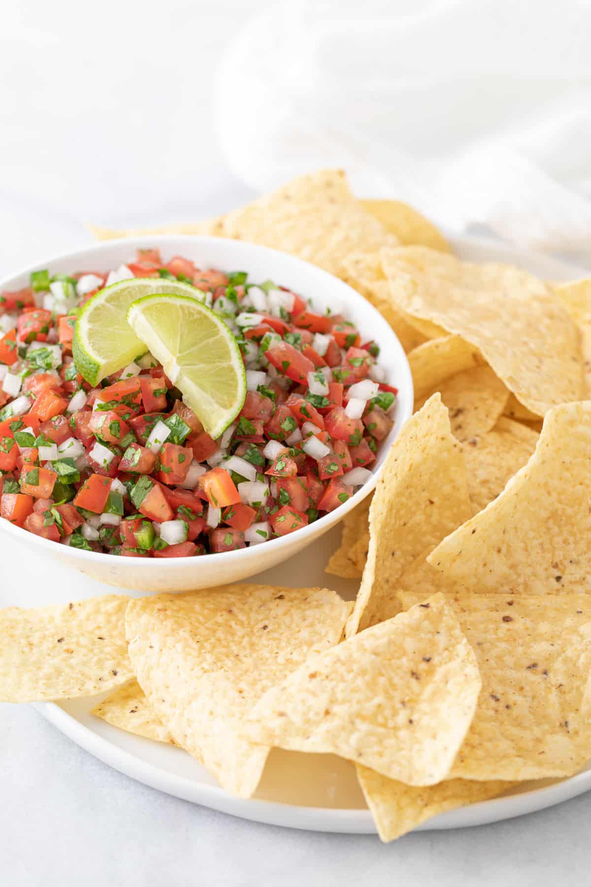 Pico de Gallo in a white bowl on a plate with tortilla chips.