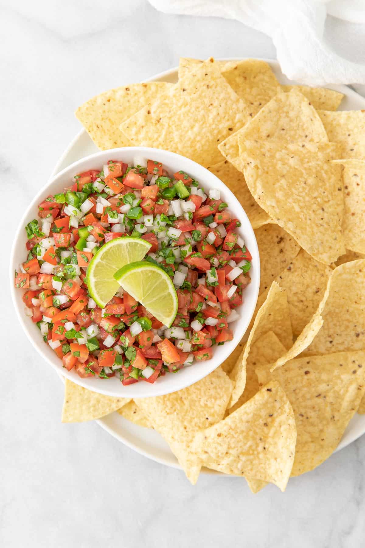 Overhead view of Pico de Gallo in a white bowl on a round plate with tortilla chips.