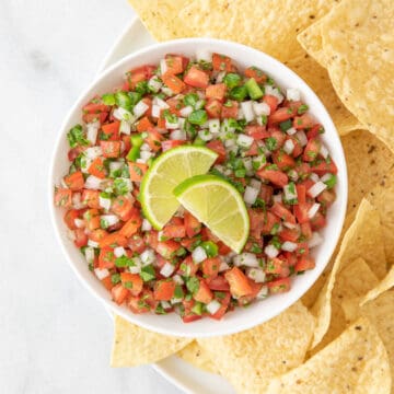 Pico de Gallo garnished with sliced limes in a white bowl on a plate with tortilla chips.