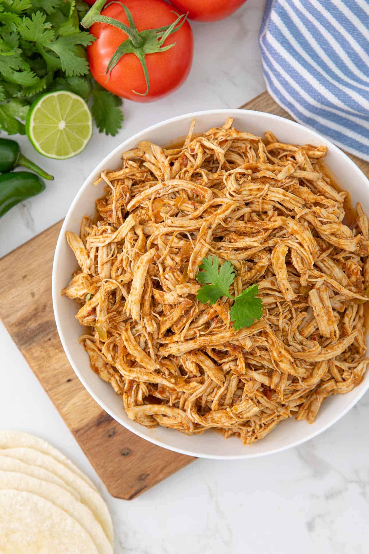 Overhead view of instant pot Mexican shredded chicken in a white serving bowl by a blue striped kitchen towel.
