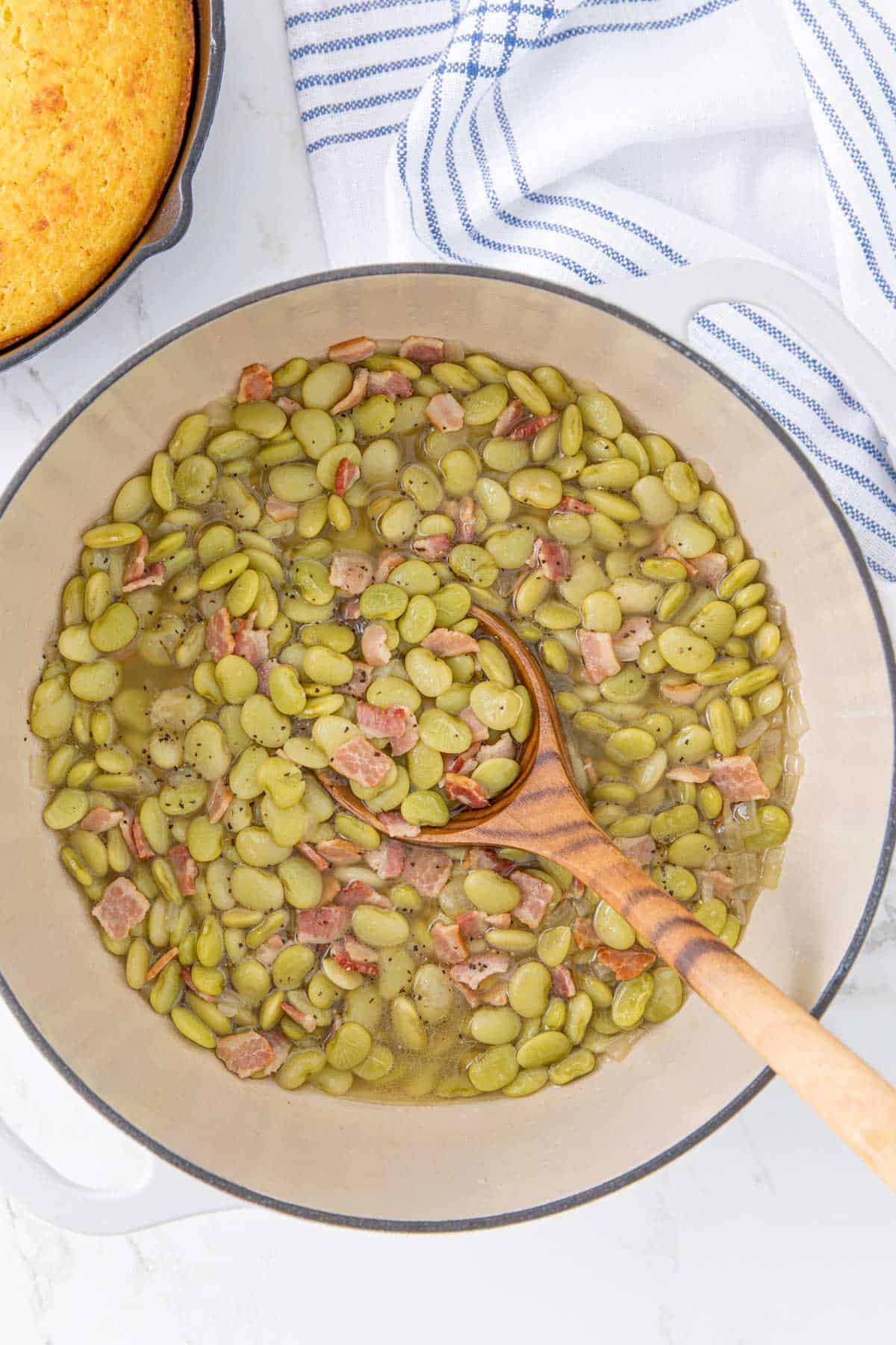 Butter beans with bacon in a white Dutch oven with a wooden ladle.