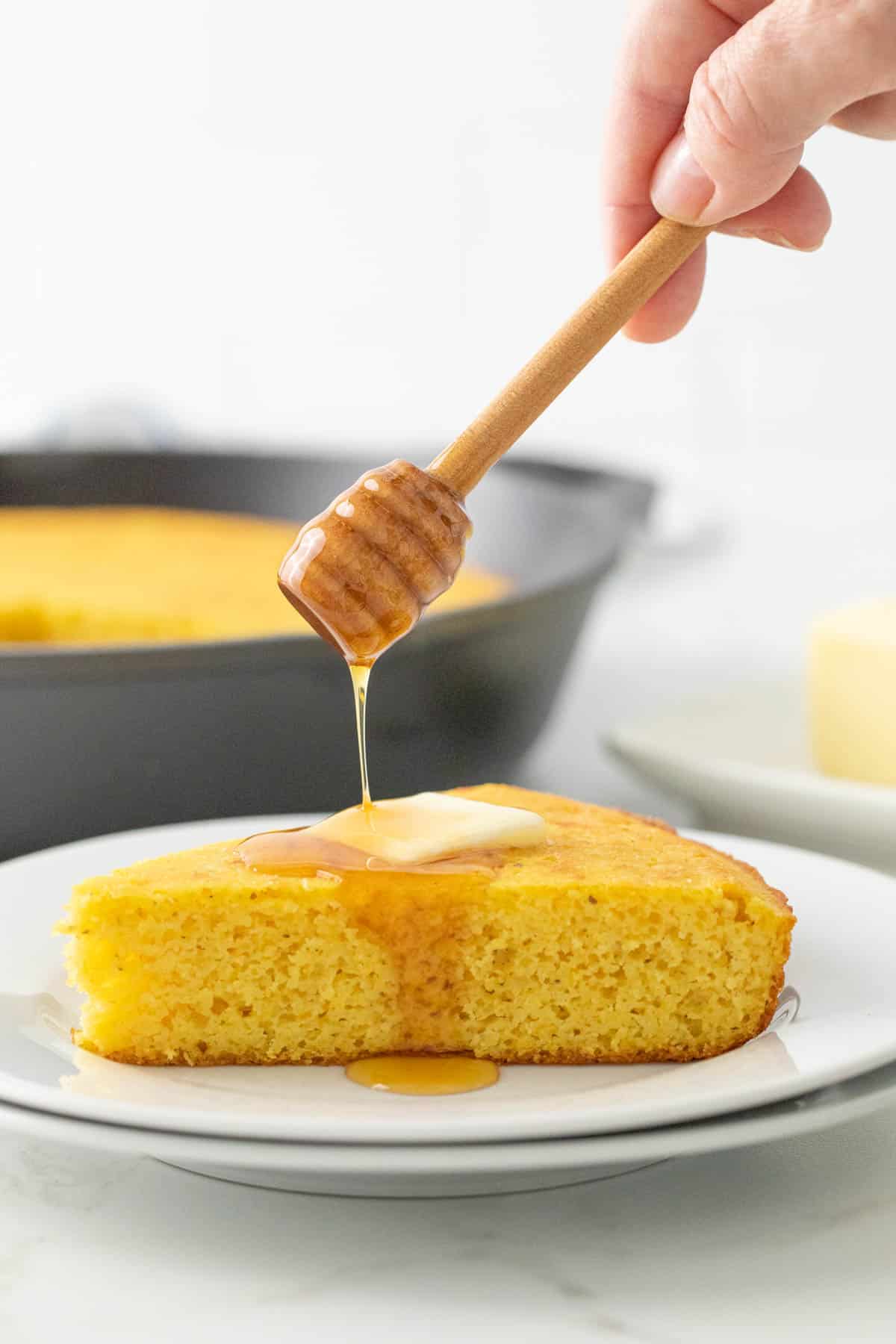 A honey dipper being held over a slice of cornbread, drizzling honey over the cornbread.