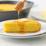A honey dipper being held over a slice of skillet cornbread, drizzling honey over the cornbread.
