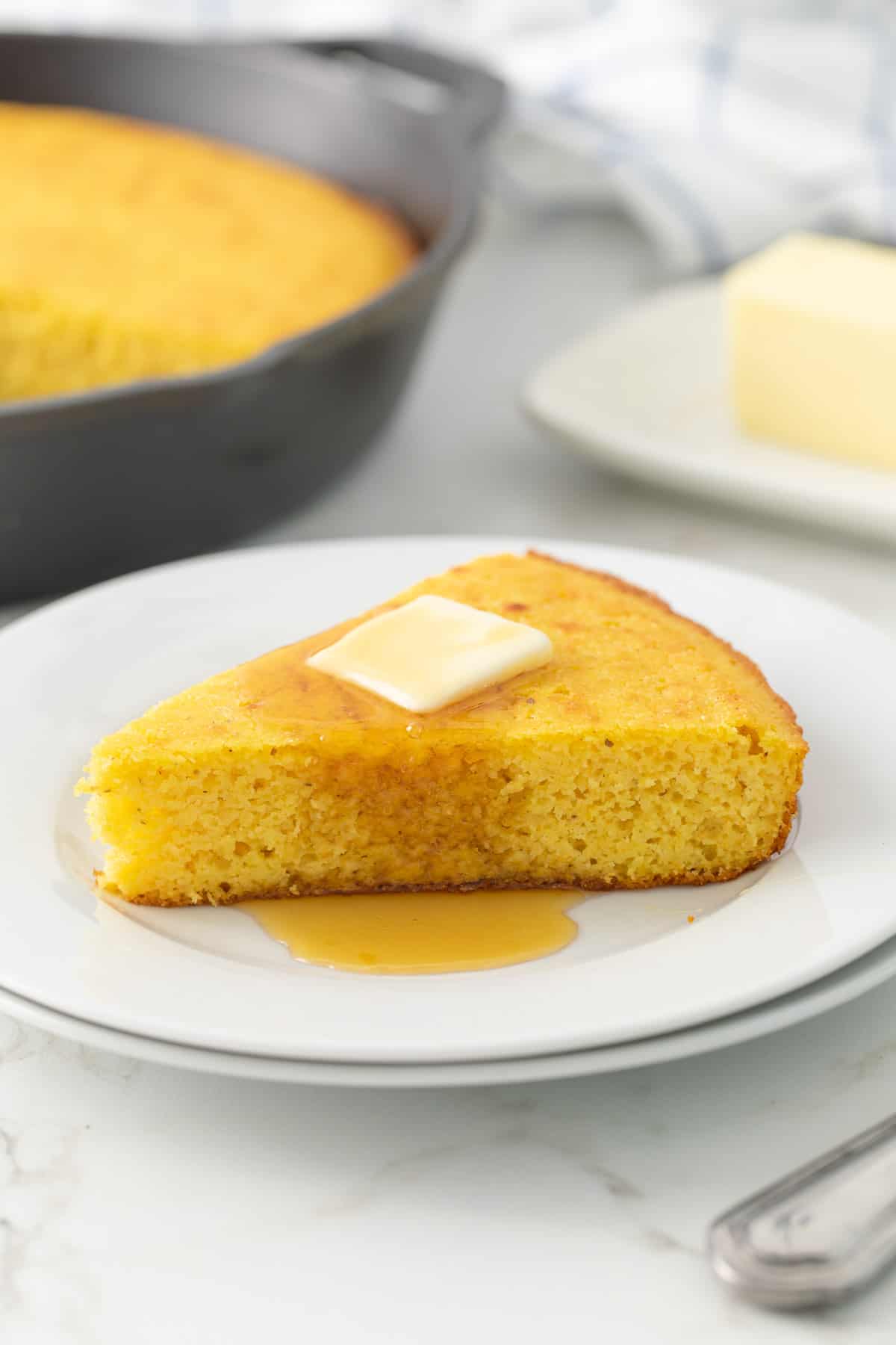 Front view of a slice of cornbread that is topped with a pat of butter and drizzled with honey on a white plate.