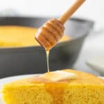 A honey dipper being held over a slice of cornbread drizzling honey on the cornbread. Overlay text at top of image.
