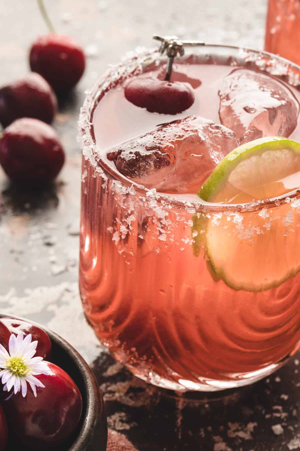 Front close-up view of a cherry margarita in a salt-rimmed glass garnished with lime and a cherry.